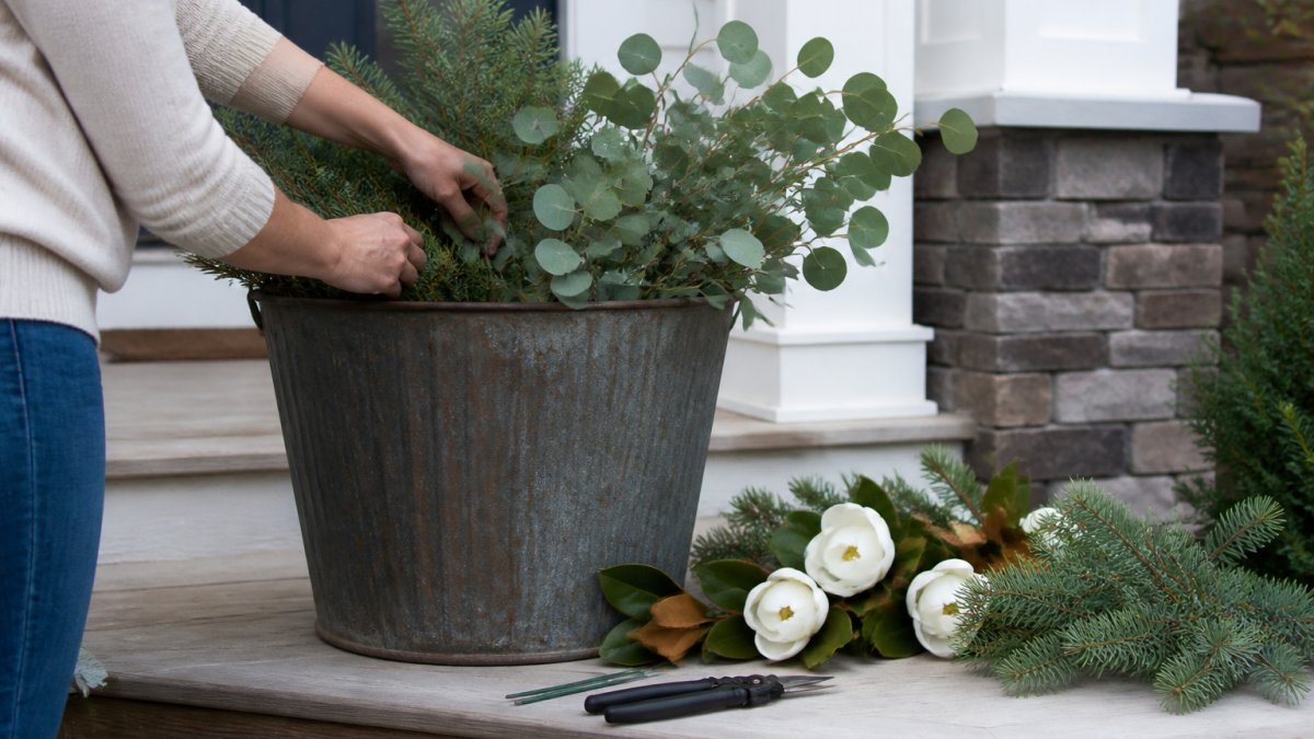 Hands placing faux greenery stems into a large rustic metal outdoor planter on a front porch, with loose magnolia blooms and pine sprigs arranged nearby on a wooden step