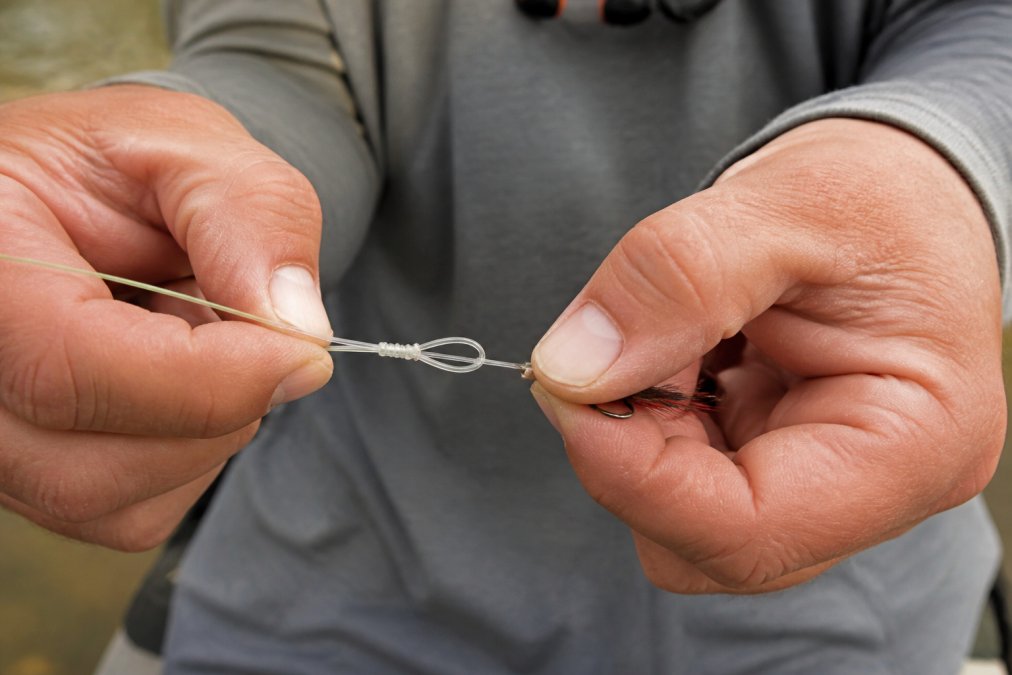 Close-up of angler's hands tying an improved clinch knot on a fly hook, showing the wrapped tag end being threaded through the loop with tippet and fly line clearly visible