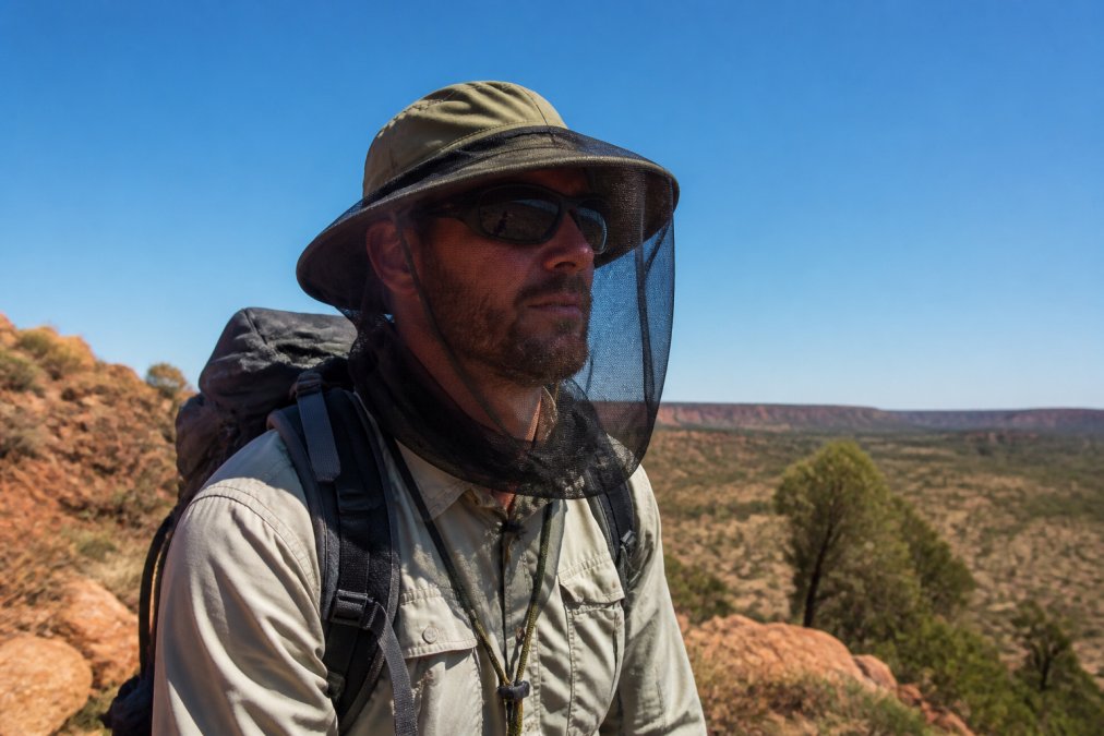 Hiker wearing a wide-brim fly net hat on a remote outback trail in harsh sunlight, with fine mesh clearly visible and the wearer's face protected from insects while navigating rocky terrain