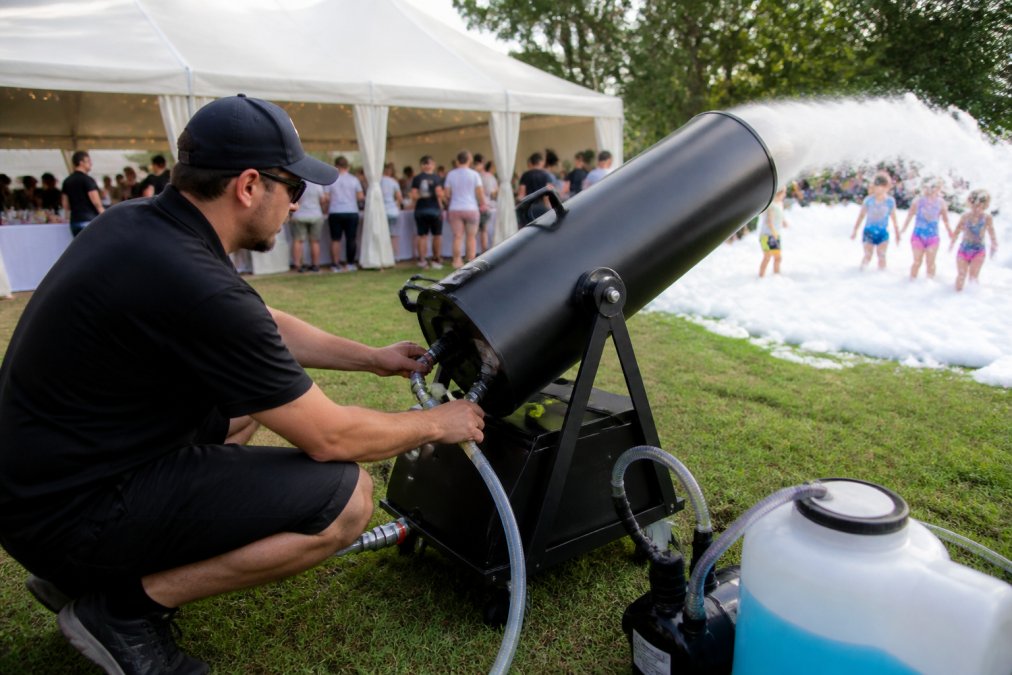 Professional demonstrating foam machine setup at outdoor party venue with water source connected and foam cannon positioned, showing equipment ready for foam dispensing with guests visible in background