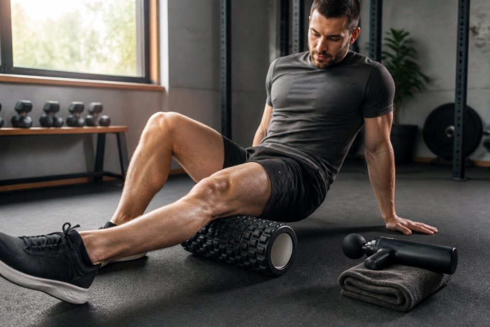 An adult athlete sitting on a gym floor rolling a foam roller slowly along their quadriceps, with a massage gun resting on a towel nearby and soft natural window light