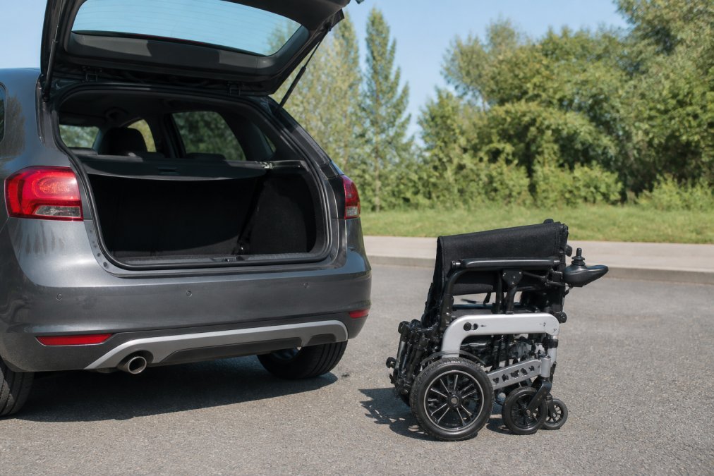 A modern lightweight folding electric wheelchair parked next to a small car with the boot open, showing how compact it is for transport