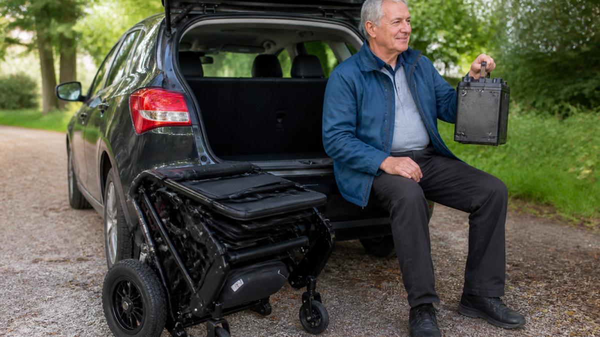 Senior adult holding a removable battery beside a folded electric wheelchair near a car trunk