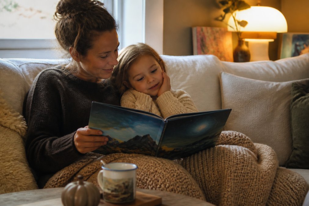 En voksen som leser høyt for barn på en koselig sofa eller lesekrok, med boken åpen mellom dem. Varmt lys fra et vindu eller lampe, barnets oppmerksomhet rettet mot boken og den voksne.