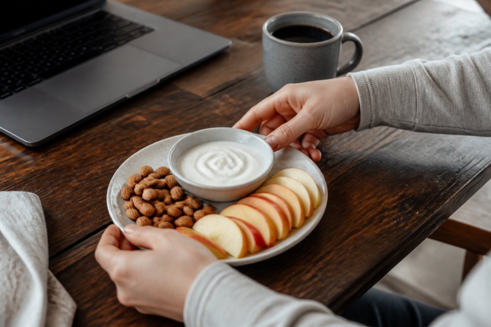 Hands arranging almonds, apple slices, and Greek yogurt for sustained energy at a workspace with a laptop.