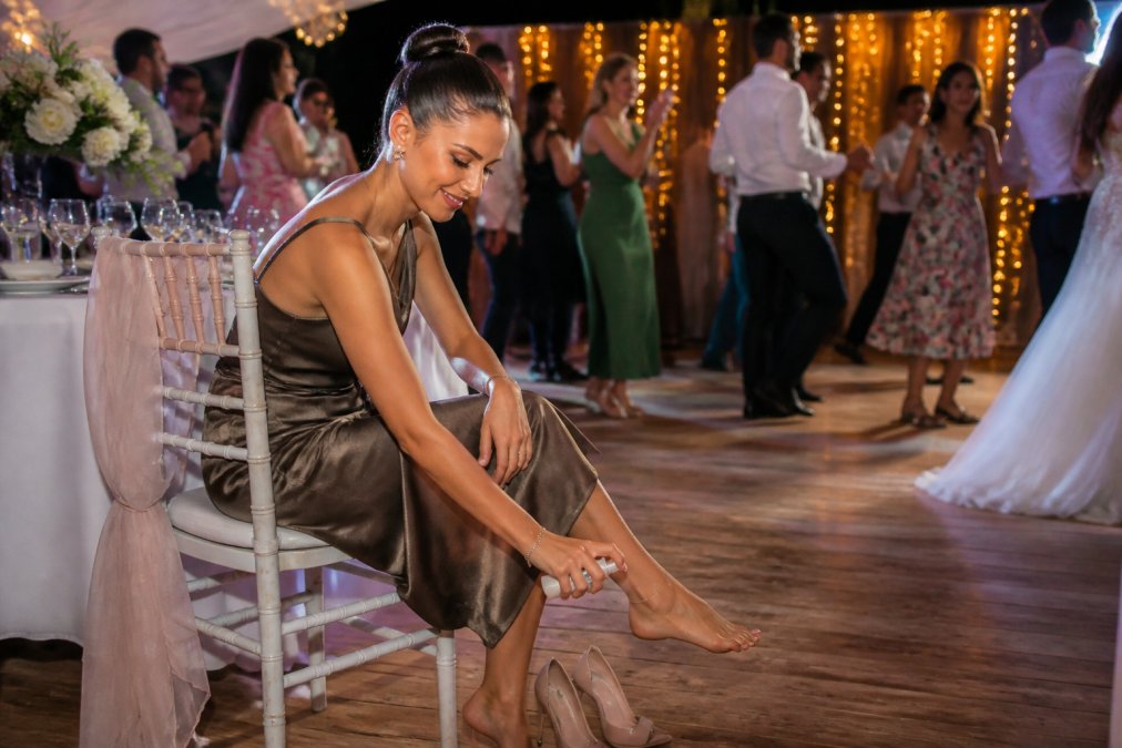 A woman in elegant high heels sitting at a wedding reception, discreetly spraying her feet with a small portable foot comfort spray bottle, warm celebratory lighting, guests dancing in the background