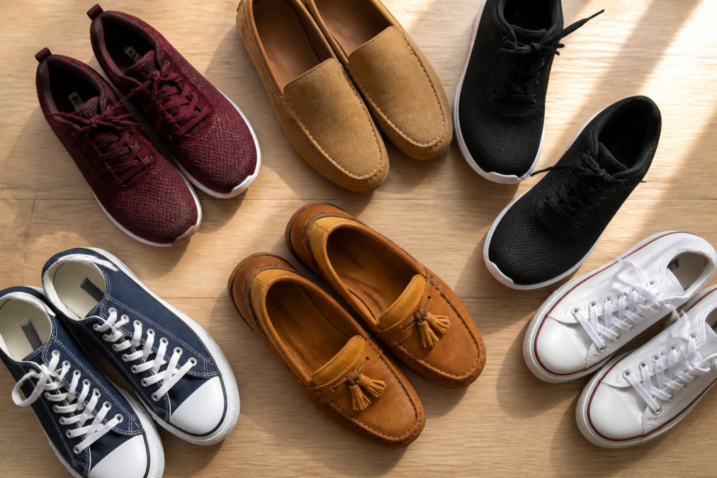 Flat lay of several pairs of shoes arranged neatly on a light wooden surface, including sneakers, loafers, and canvas shoes in various colors, photographed in warm natural light