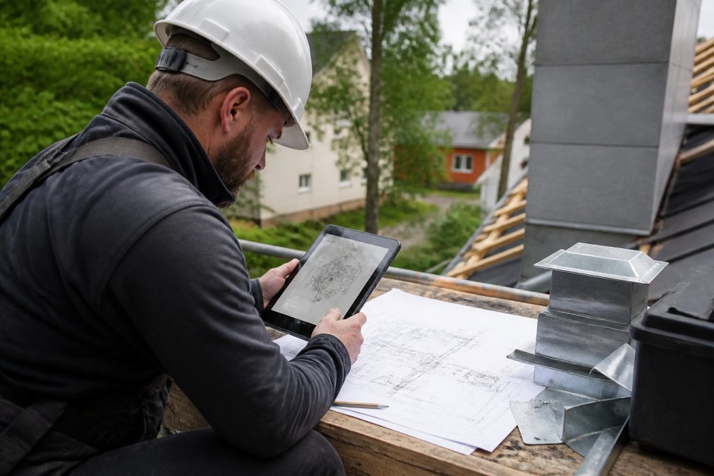 Håndverker på byggeplass som bruker tablet for å bestille beslag, med tekniske tegninger og målinger synlige på skjermen og papirer rundt ham