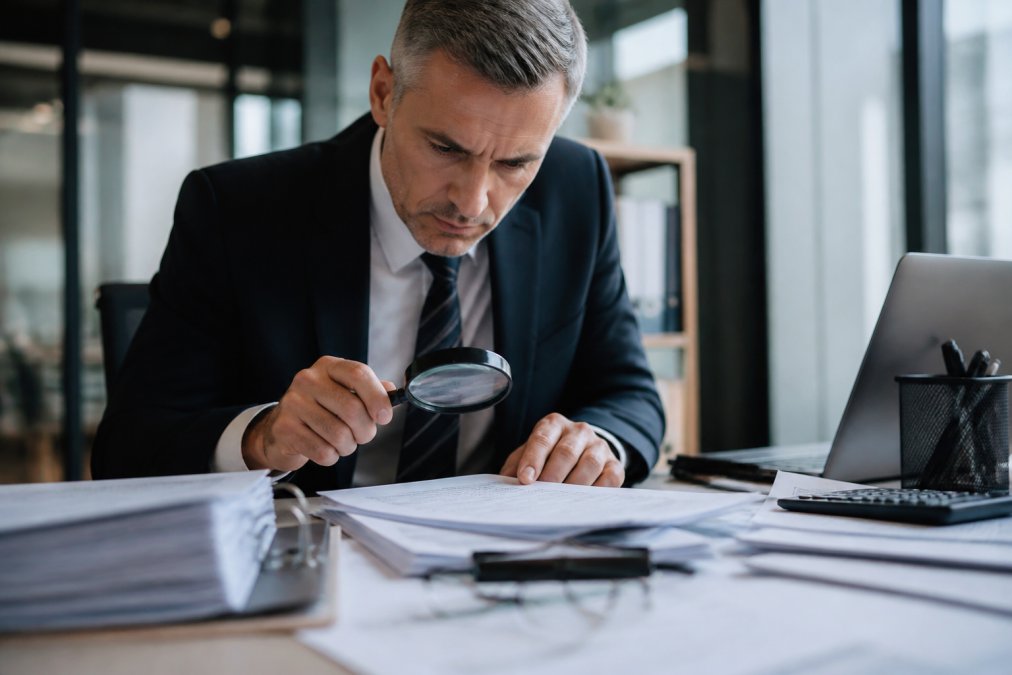 A forensic accountant examining financial documents with a magnifying glass in a modern office, with a laptop showing spreadsheets in the background