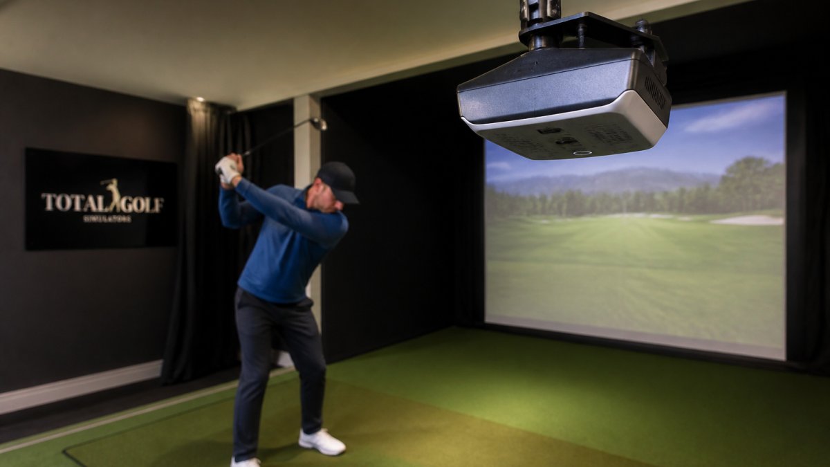 Close-up of a launch monitor mounted above a golf hitting mat in a home simulator setup, with a golfer mid-swing in the background, showing the device's position relative to the hitting area and projection screen