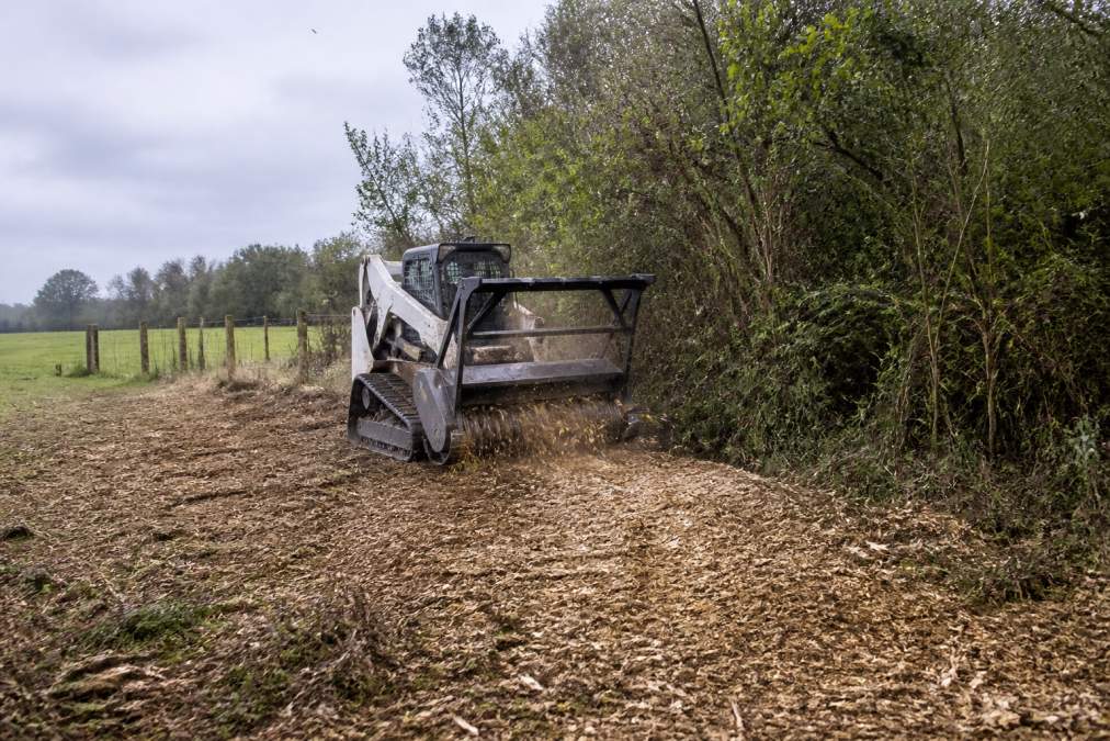 A forestry mulcher attachment on a tracked skid steer grinding thick brush and small trees along a wire fence line on a rural pasture, fresh wood mulch spread on the ground behind the machine, overcast morning light