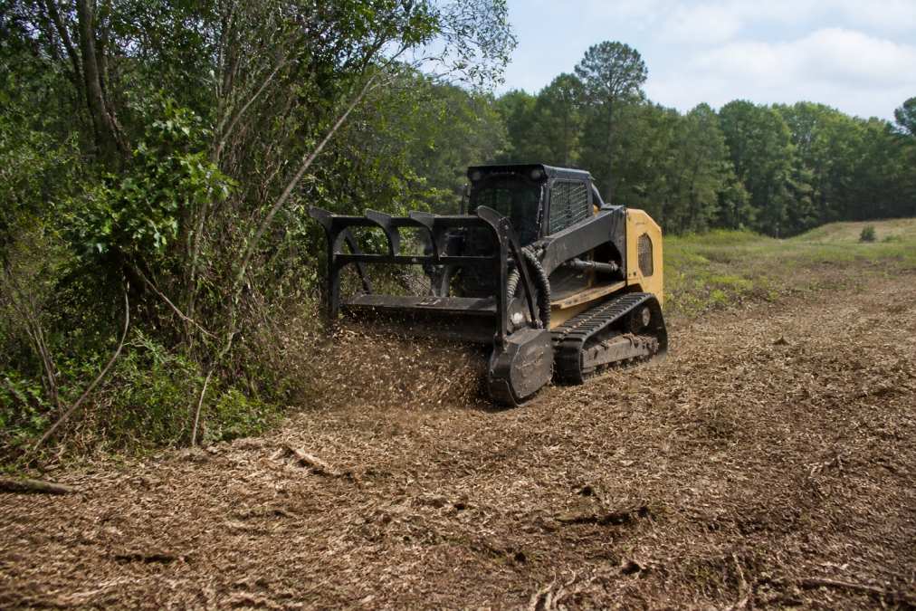 A tracked forestry mulcher grinding through dense brush and small trees in an East Texas pasture, with a fine layer of wood mulch spreading across the cleared ground behind it