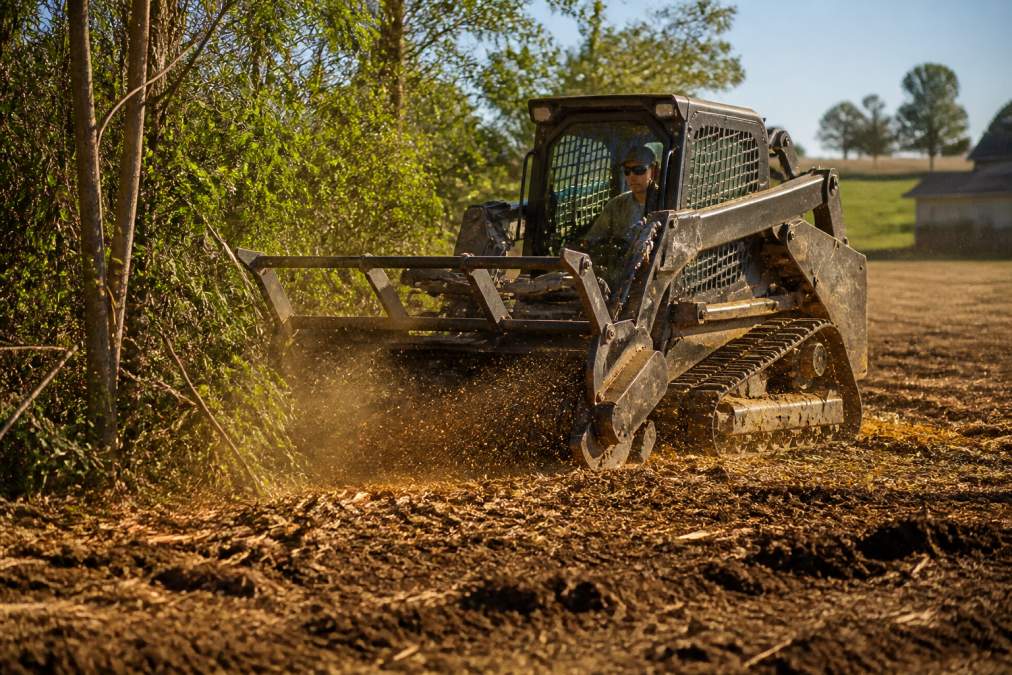 A tracked forestry mulcher grinding brush and small trees into fine mulch on a rural property, operator visible in the cab, golden afternoon light