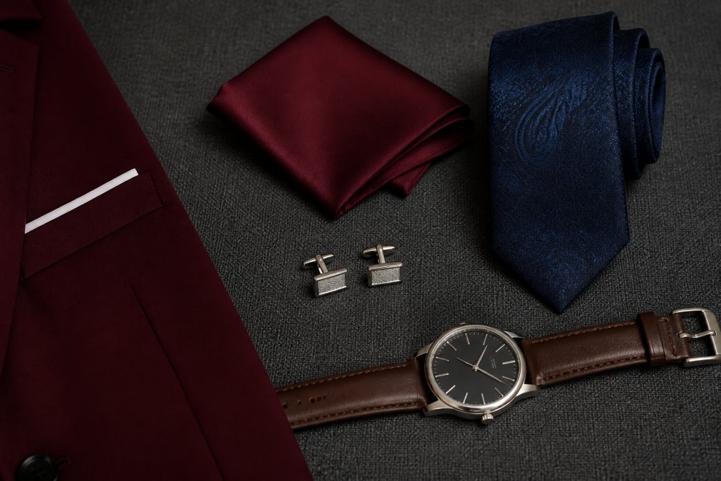 Flat lay of formal business accessories including silver cufflinks, burgundy pocket square, navy silk tie, and leather dress watch arranged neatly on dark charcoal fabric next to suit material