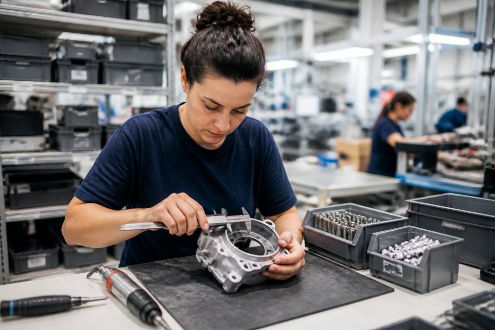 An operator in a clean industrial workshop performing a quality check on an assembled mechanical component, using precision tools on a workbench with organized parts bins nearby