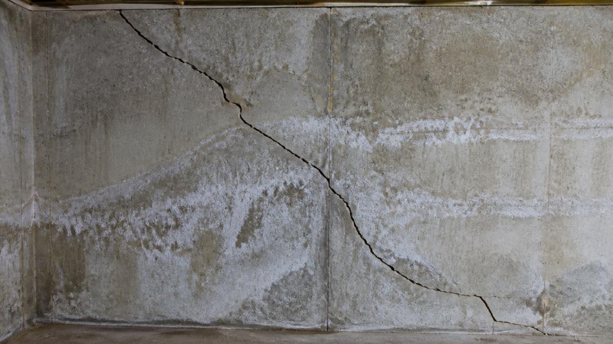 Close-up of basement concrete wall showing visible diagonal foundation cracks, water staining, and efflorescence deposits on the surface under bright inspection lighting