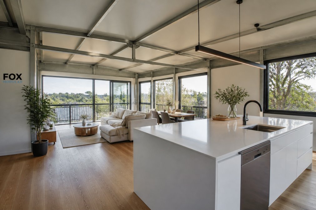 Interior view of a modern modular home with open-plan living area, steel-framed structural elements visible, natural light streaming through large windows, and contemporary furnishings in a bright, spacious room