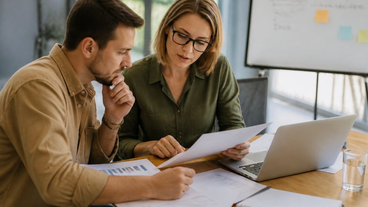 A business owner and a financial consultant reviewing printed financial statements and a laptop at a table in a bright office, with a whiteboard of growth targets behind them