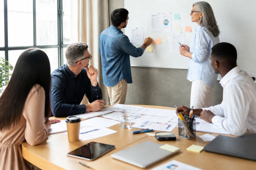 Diverse marketing team collaborating in a modern office, reviewing strategy documents and discussing implementation on a whiteboard, bright natural lighting