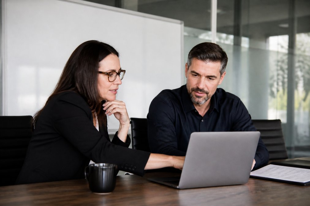 A small business owner and a fractional executive reviewing an operations dashboard together at a conference table, laptops open, whiteboard with workflow notes behind them
