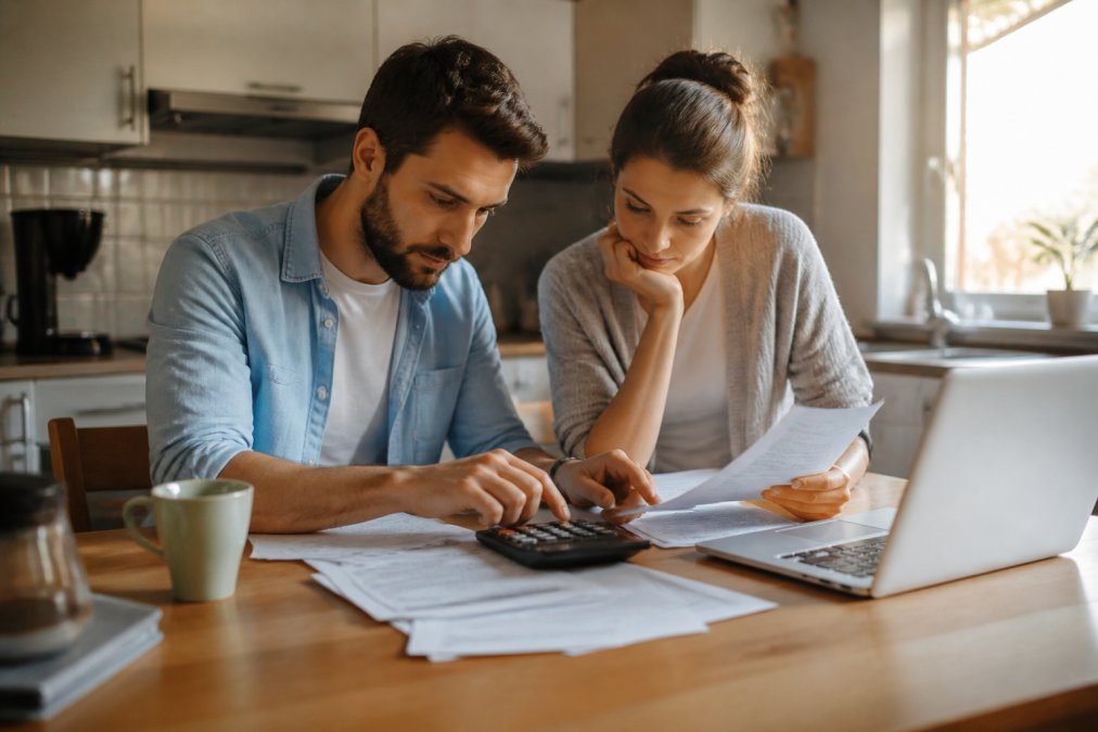 Un couple assis à une table de cuisine avec des documents étalés, une calculatrice et un ordinateur portable ouvert, examinant leur budget d'achat sous une lumière naturelle de fin d'après-midi