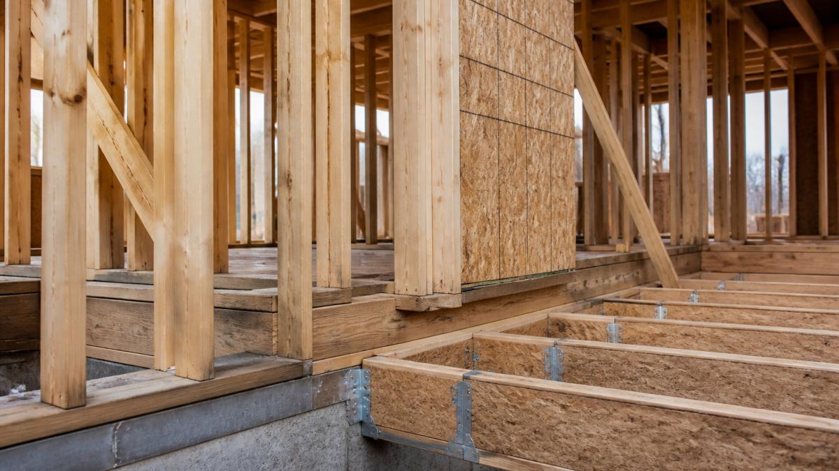 Close-up of structural framing components on an active job site, showing wall studs, floor joists, metal connectors, and fasteners during mid-construction, with natural daylight illuminating the exposed frame