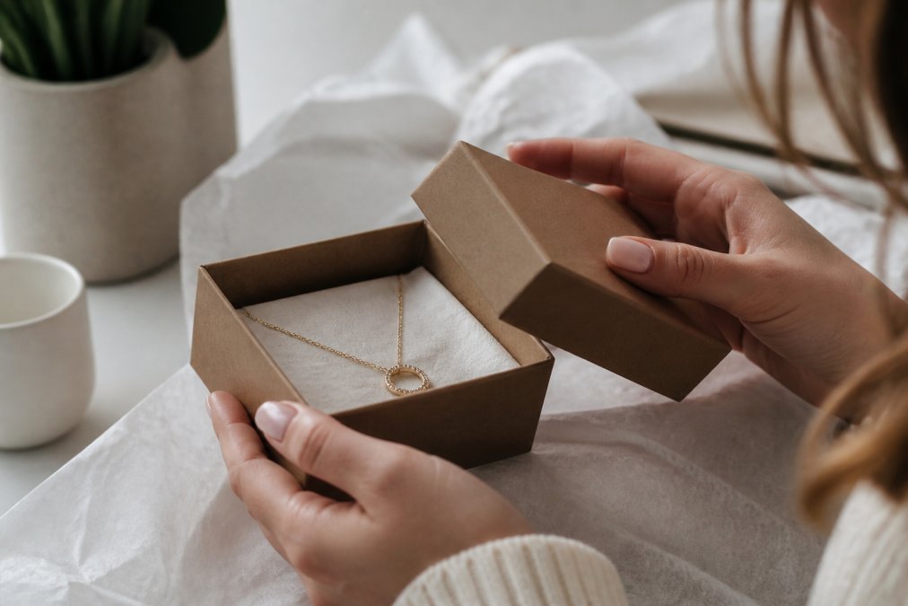 A person's hands carefully lifting a small rigid gift box lid, revealing a delicate gold necklace nestled in soft tissue paper, warm natural light falling across a clean white table