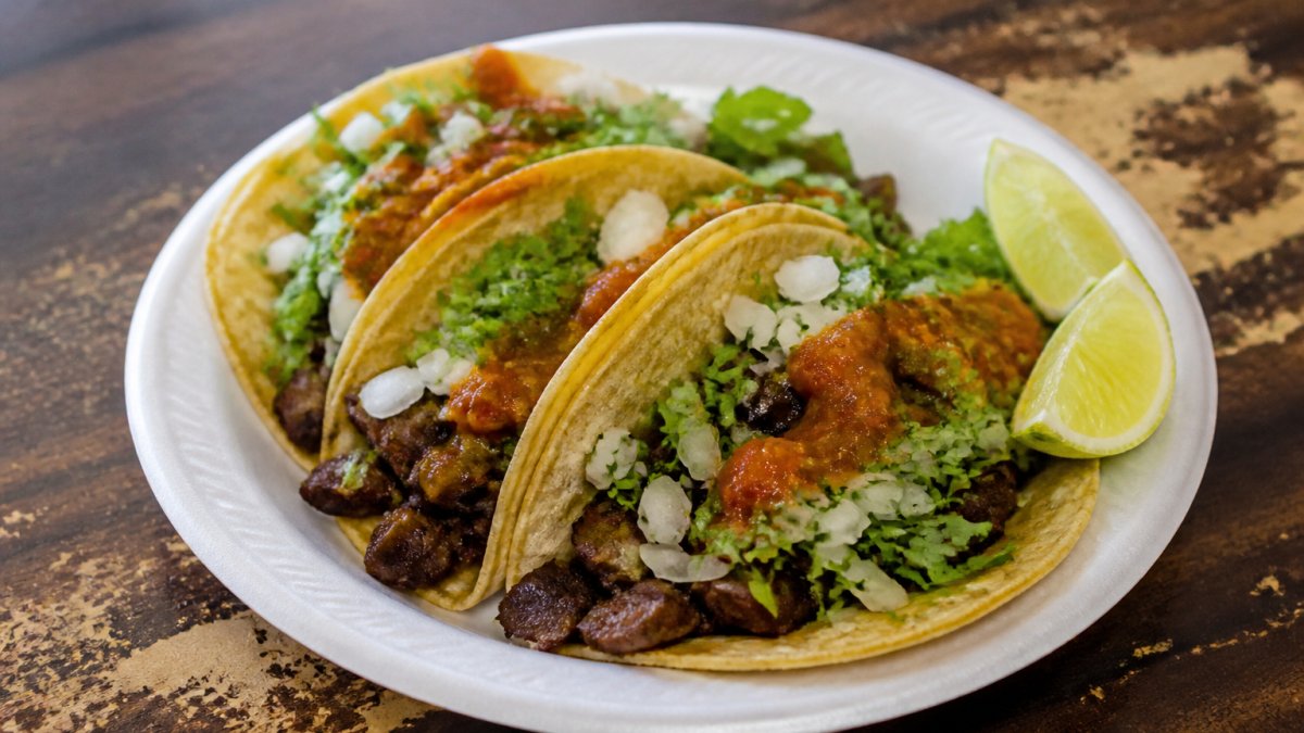 Close-up of three street tacos on a paper plate with lime wedges, chopped onion, cilantro, and salsa roja, served on a worn wooden table at a casual taquería