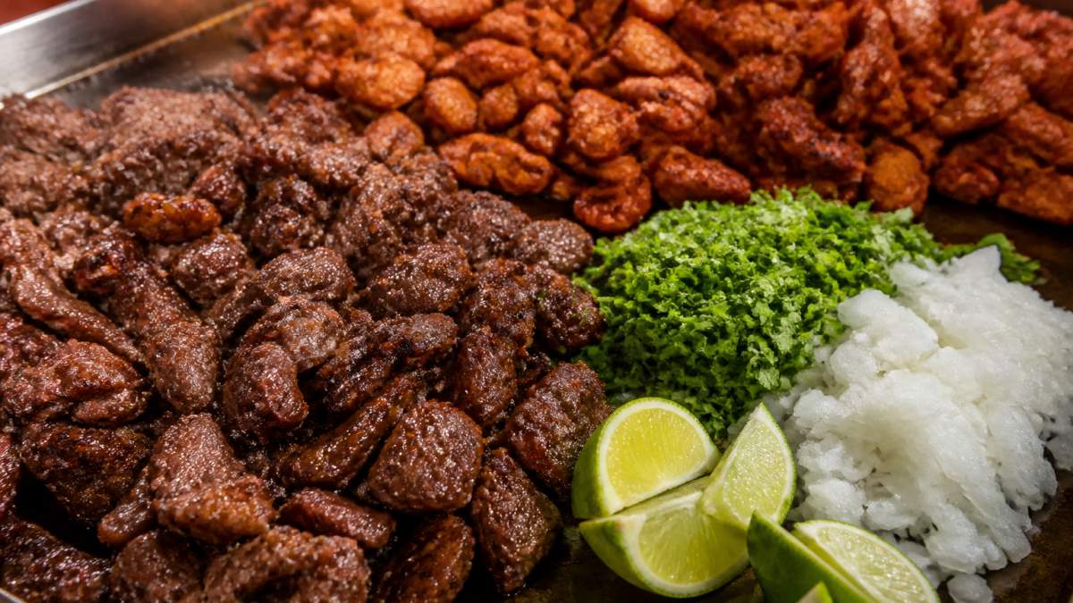 Close-up of freshly grilled carne asada and pastor meats sizzling on a hot griddle, with vibrant cilantro, diced white onions, and fresh lime wedges arranged alongside