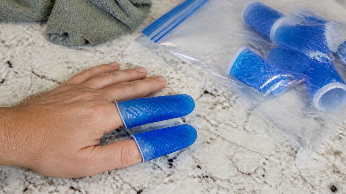 Close-up of an adult's hand with a stretchy blue gel compression sleeve fitted over the index and middle fingers, resting on a kitchen counter next to a freezer bag of ice packs
