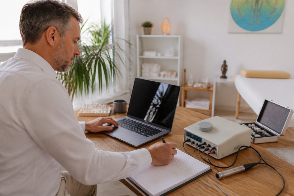 A Heilpraktiker sitting at a tidy wooden desk in a bright treatment room, operating a radionics software interface on a laptop, with a frequency therapy device placed beside the screen and a notebook with handwritten session notes