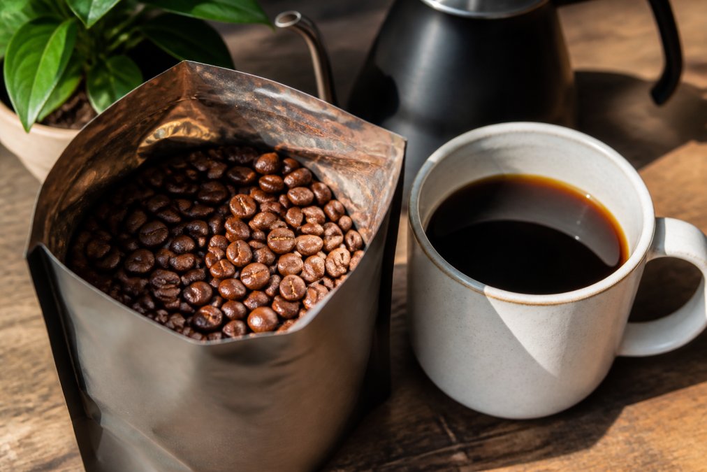 Close-up of fresh whole coffee beans in an open bag with glossy surfaces reflecting light, displayed next to a cup of brewed coffee in natural morning sunlight