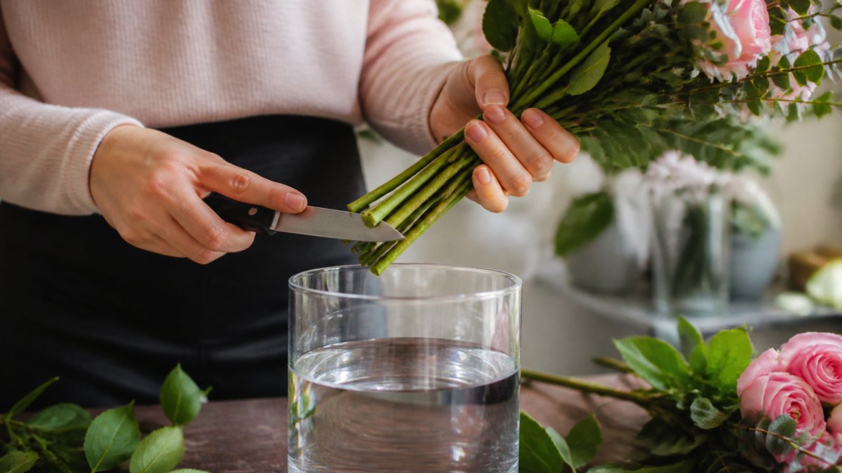 Close-up of hands holding fresh cut flowers and a sharp knife, trimming stems at a 45-degree angle over a clean vase with water
