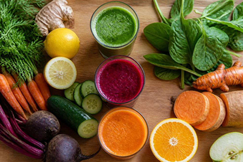 Overhead shot of three freshly pressed juices in clear glasses displaying vibrant green, deep red, and orange colors, with fresh vegetables and fruits arranged beside them on a wooden counter in natural morning light