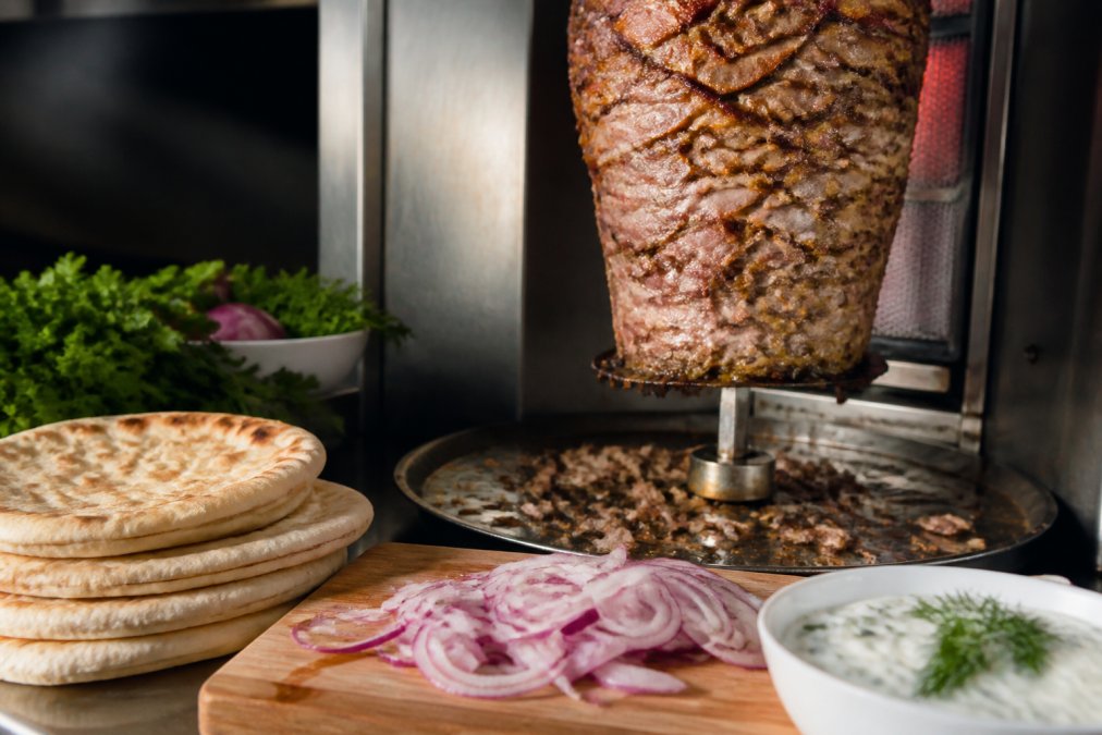 Close-up of marinated lamb meat slowly rotating on a vertical rotisserie spit with golden-brown exterior, fresh pita bread stacked nearby, bright red onions sliced on a cutting board, and a white bowl of creamy tzatziki sauce with herbs, natural kitchen lighting