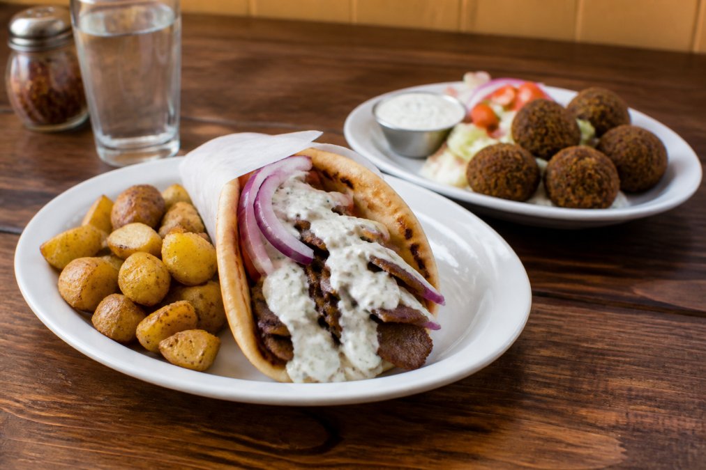 Fully plated lamb gyro with generous portions of marinated meat, warm pita bread folded around filling, creamy tzatziki sauce drizzled on top, fresh red onions visible, accompanied by golden lemon potatoes on one side and crispy falafel pieces on the other plate, warm restaurant lighting