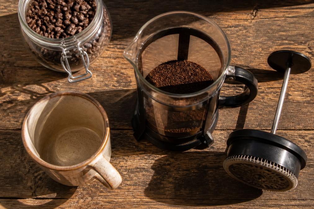 Overhead view of a French press filled with fresh coffee grounds, ceramic mug nearby, and whole bean coffee in a storage container on a rustic wooden table with morning light