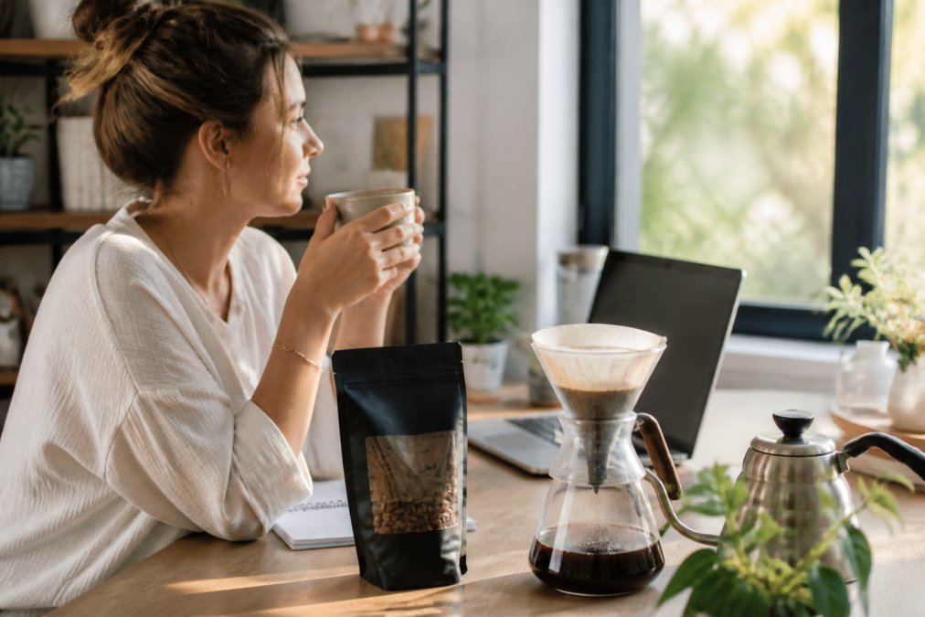A remote worker sitting at a bright home office desk with a pour-over coffee setup, a sealed bag of whole bean coffee with a visible roast date, and an open laptop in front of a large window with natural morning light