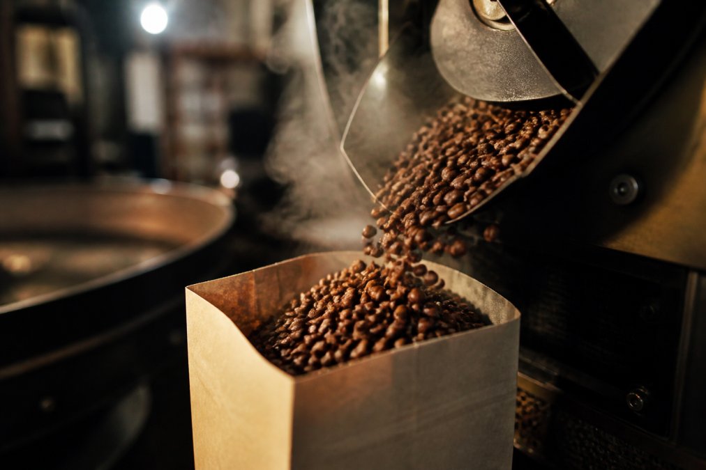 Close-up of freshly roasted coffee beans being poured into a kraft paper bag in a roastery, with steam rising and warm lighting highlighting the rich brown color and glossy texture of the beans