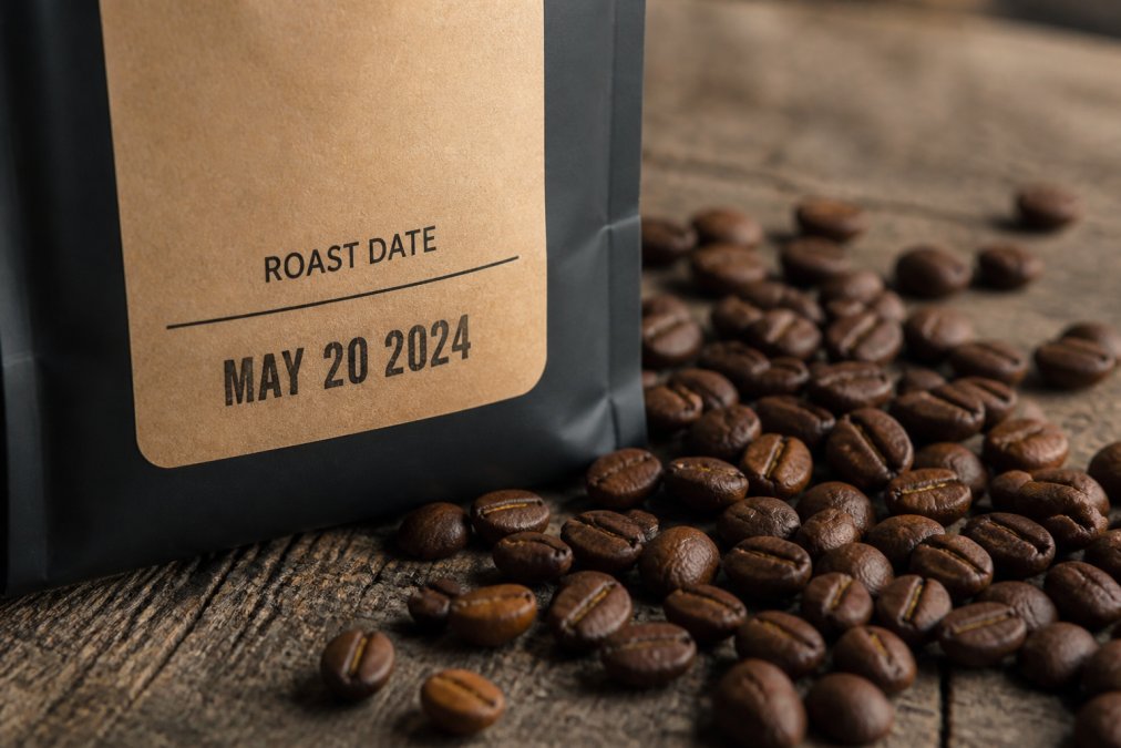 Close-up of a coffee bag label displaying the roast date clearly printed in bold text, with whole roasted beans scattered on a weathered wooden surface next to the bag
