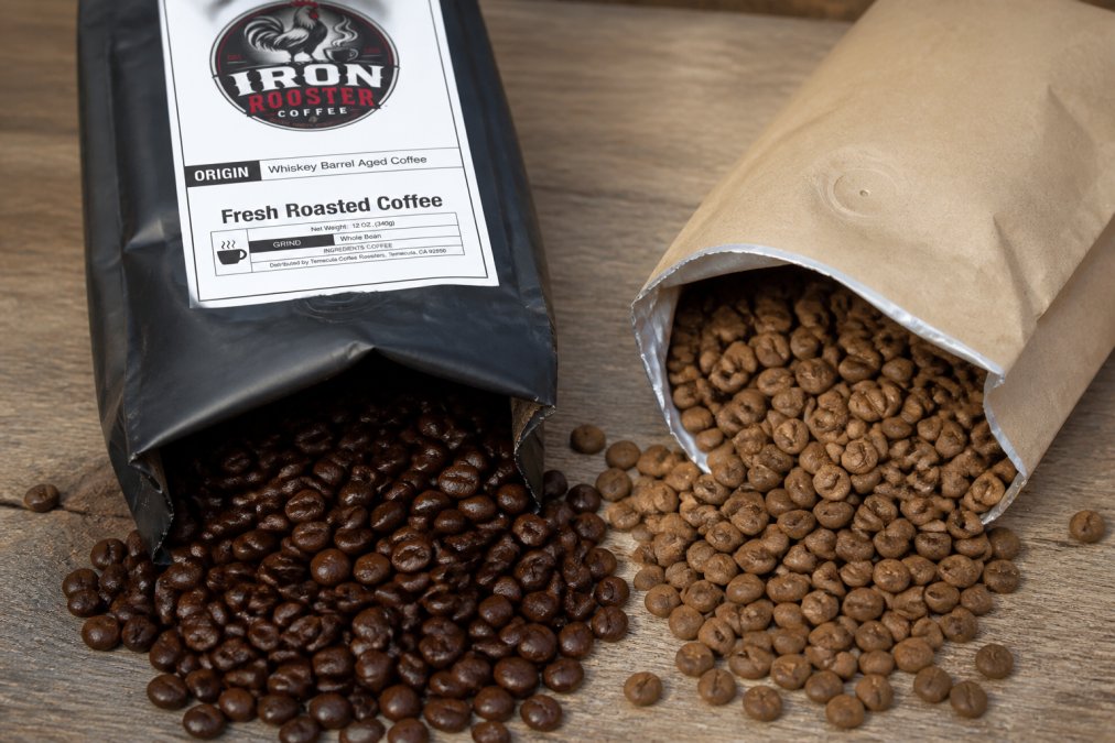 Close-up of freshly roasted dark brown coffee beans in a valve-sealed bag next to an open bag of faded, dull grocery store coffee beans, showing the stark color and texture difference in natural light