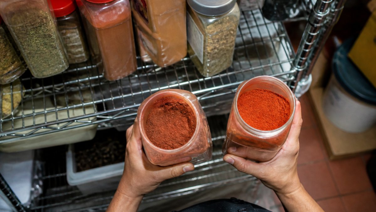 Overhead shot of a restaurant dry storage shelf with dated spice containers, one jar open showing faded reddish powder next to a fresh vivid jar, fluorescent lighting