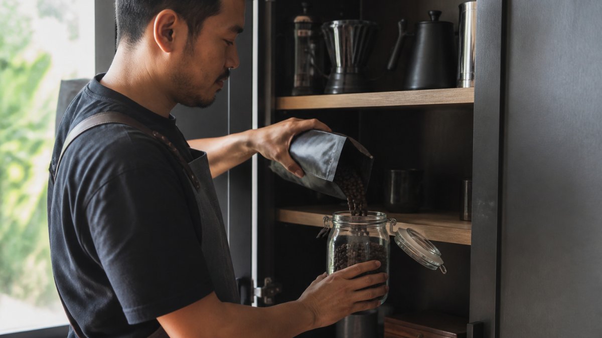Professional barista pouring freshly roasted whole coffee beans into a glass airtight container with a sealed lid, stored in a cool dark kitchen cabinet with natural morning light