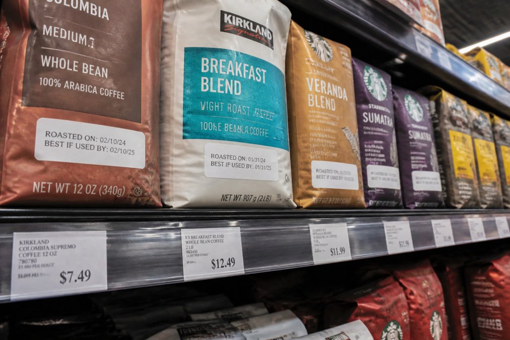 Close-up of multiple coffee bags displayed on a grocery store shelf with various roast date labels and brand names visible on packaging in bright fluorescent lighting