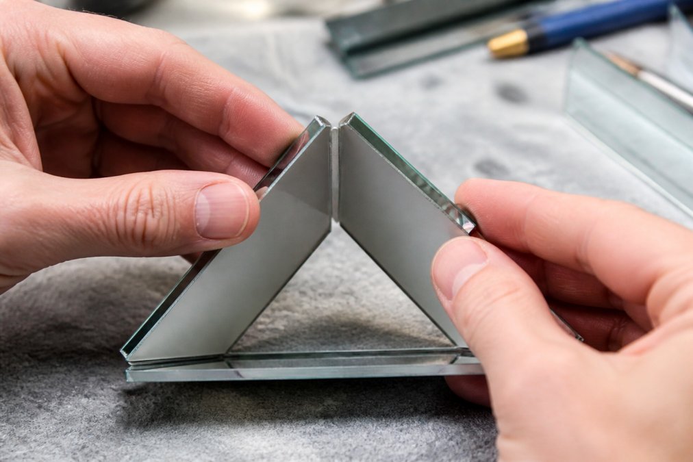 Hands carefully assembling three front surface mirror strips into a triangular tube configuration, showing precise alignment and the protective spray coating on the aluminum-coated glass surfaces under bright workshop lighting