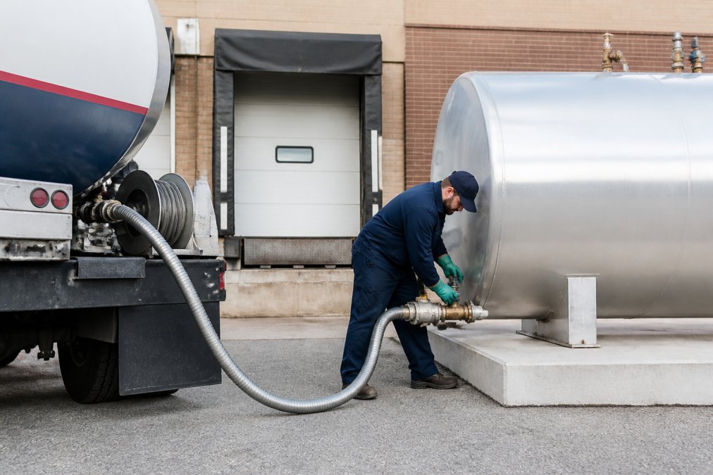 Propane delivery truck backed up to a commercial loading dock with a uniformed technician connecting a silver delivery hose to a large cylindrical storage tank mounted on concrete