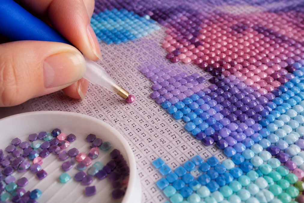Close-up of a hand holding a drill pen with round and square diamond drills visible on a canvas, showing the sparkle and facet detail of each shape against the color-coded legend