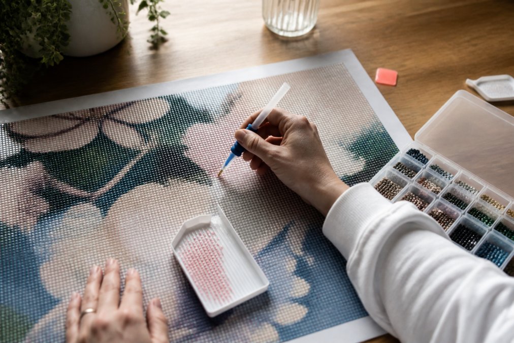 Crafter's hands working on a full drill diamond painting canvas with an applicator tool, tray of drills, and wax visible, demonstrating proper placement technique and workspace setup