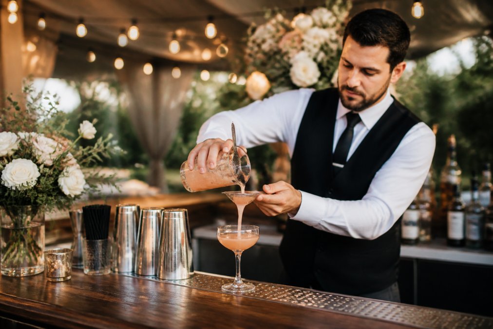 Bartender pouring a craft cocktail at an elegant wedding bar package setup with floral decor and string lights