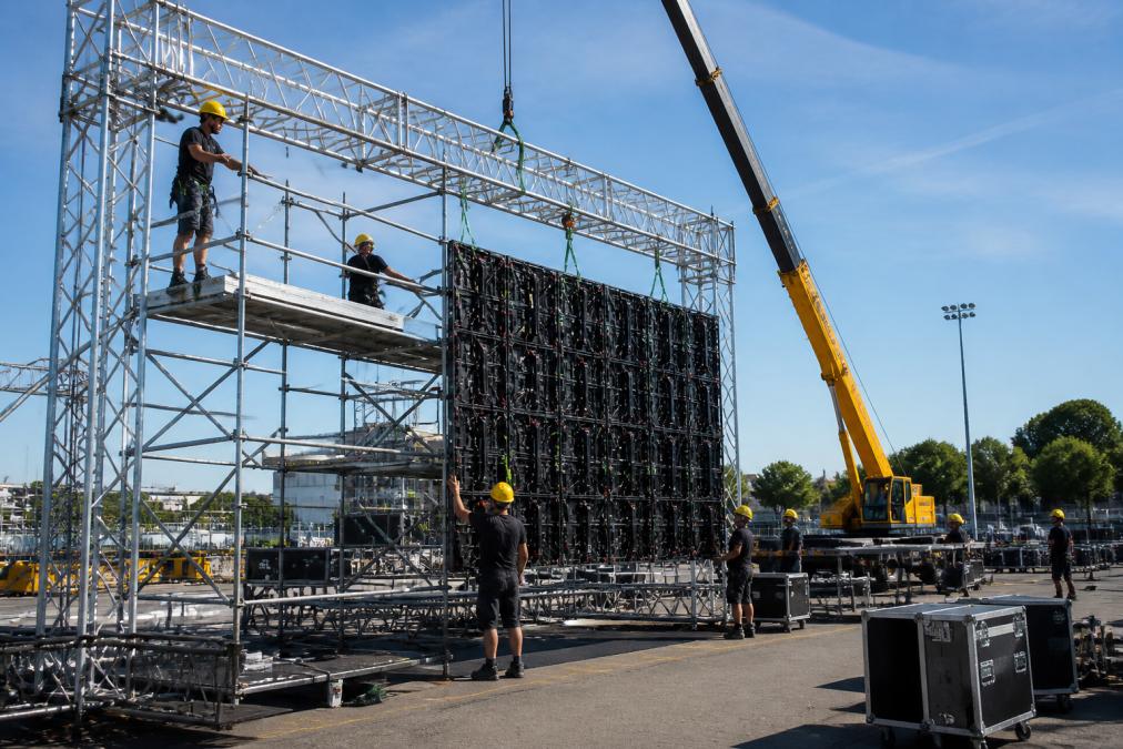 Professioneel team dat een groot LED-scherm op een evenementenlocatie opbouwt met kraan, steigers en gele veiligheidshelmen, op een zonnige dag met duidelijk zicht op modulaire panelen